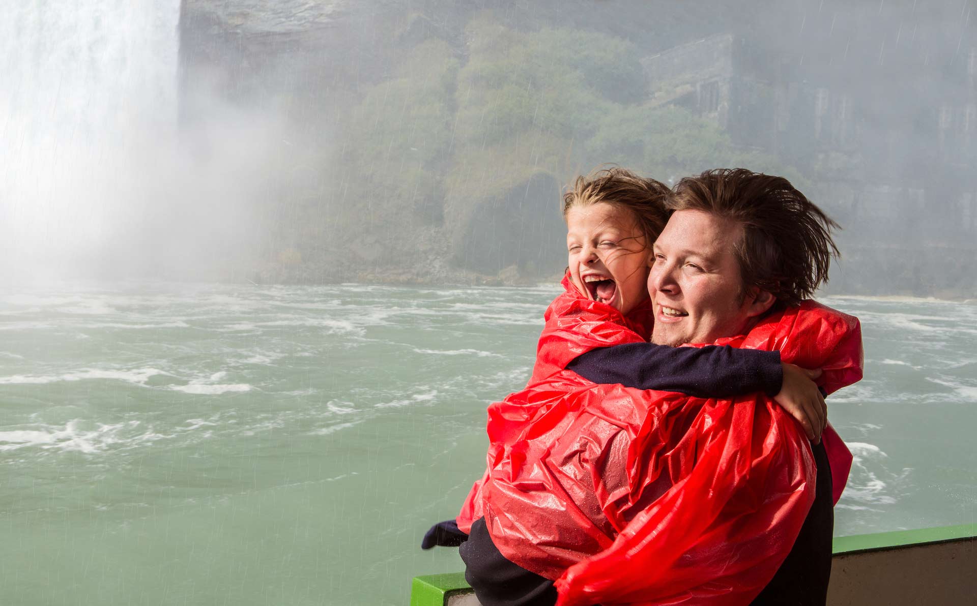 Adult holding a child on boat at base of Niagara Falls getting wet wearing ponchos.